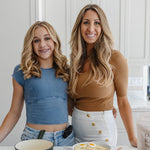 Kolbie and Janae Cox standing in a bright kitchen behind a table with food items, including a bowl of pasta with eggs, a basket of greens, and a pot.