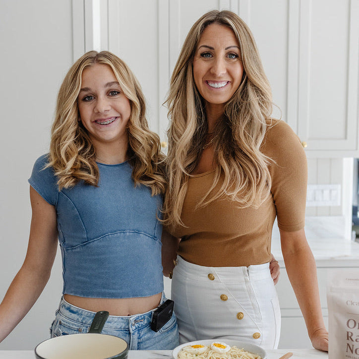 Kolbie and Janae Cox standing in a bright kitchen behind a table with food items, including a bowl of pasta with eggs, a basket of greens, and a pot.
