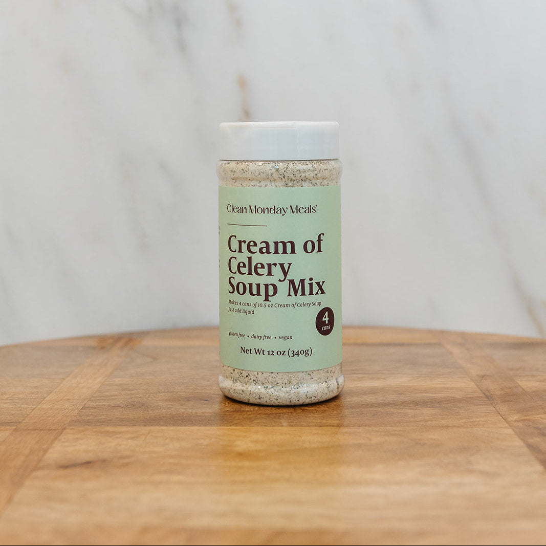 A bottle of Cream of Celery Soup Mix by Clean Monday Meals sits on a wooden cutting board against a white marble background.