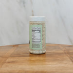 A clear plastic spice jar filled with a light beige seasoning blend, featuring a white lid and a light green label with a visible nutrition facts panel, set on a wooden surface against a white marble background.