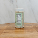 A clear plastic spice jar filled with a light beige seasoning blend, featuring a white lid and a light green label with a visible nutrition facts panel, set on a wooden surface against a white marble background.