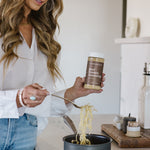 Janae Cox cooking beef ramen next to a stove and in a black pot while holding a bottle of the clean Monday Meals ramen seasoning. 