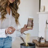 Janae Cox cooking beef ramen next to a stove and in a black pot while holding a bottle of the clean Monday Meals ramen seasoning. 