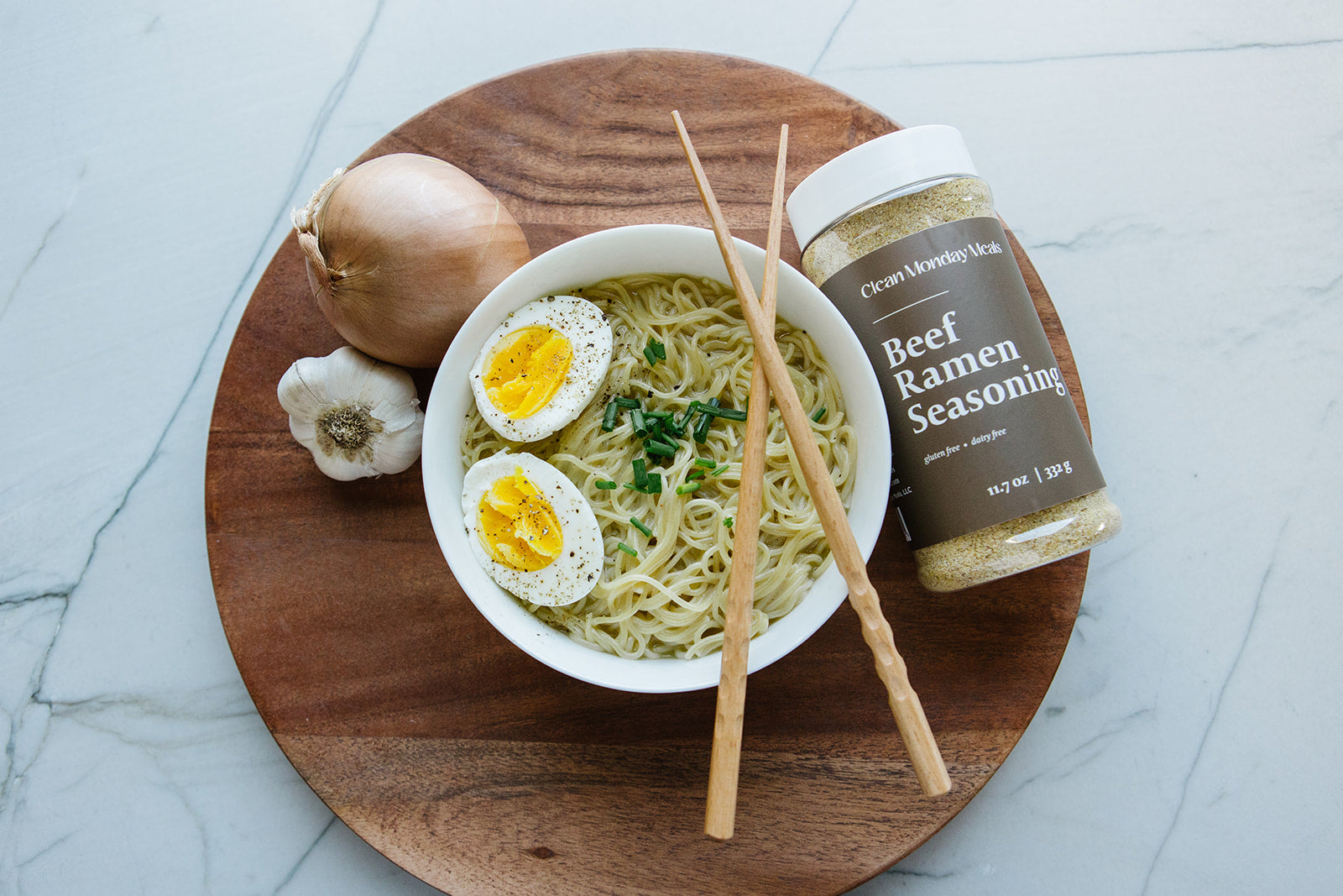 Beautiful overhead shot of Clean Monday Meals Ramen Seasoning sitting on a wood serving tray, next to a bowl of ramen noodles with an egg cut in have and seasoning with salt and pepper. 