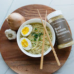 Beautiful overhead shot of Clean Monday Meals Ramen Seasoning sitting on a wood serving tray, next to a bowl of ramen noodles with an egg cut in have and seasoning with salt and pepper. 