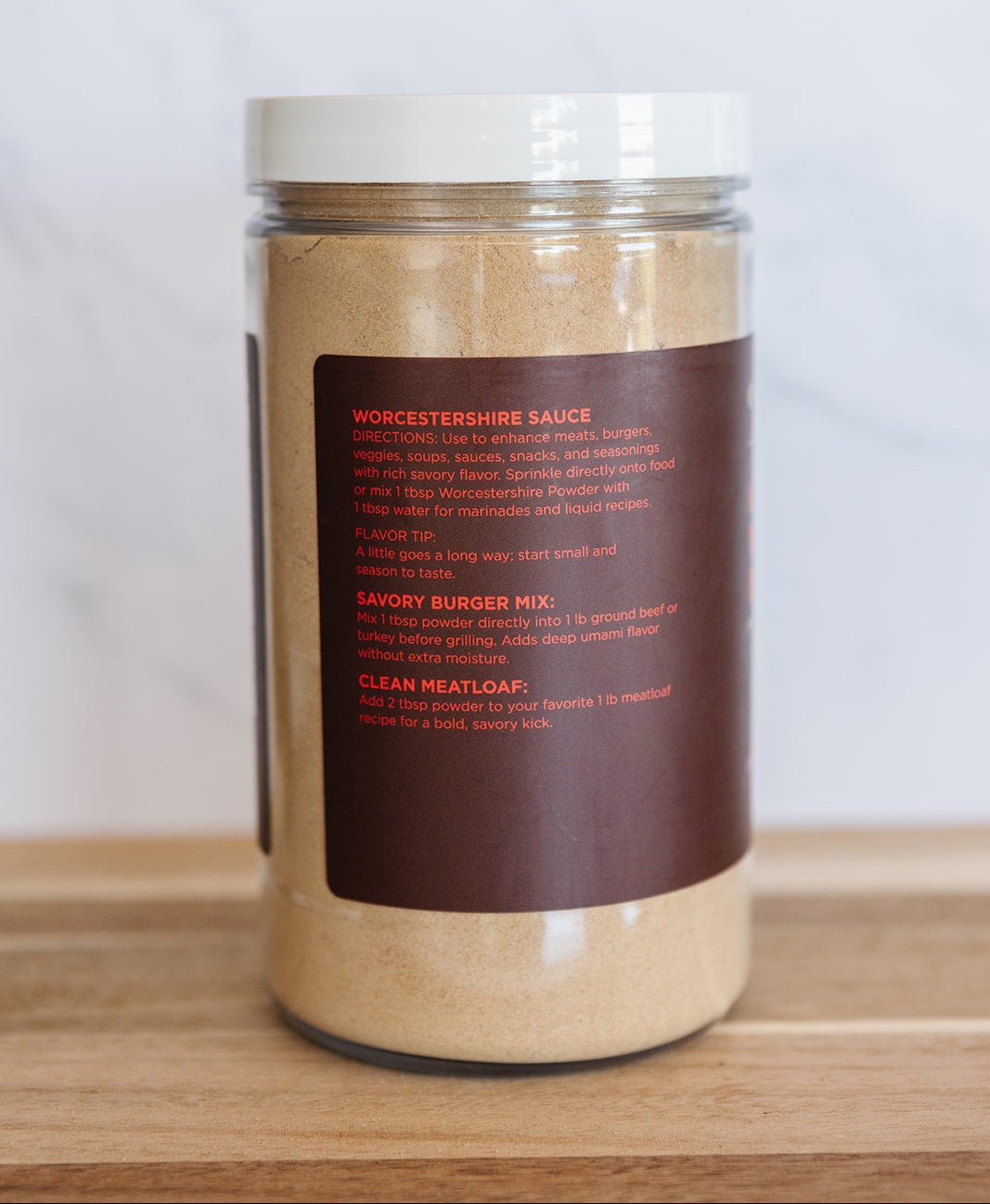 A clear plastic jar of Worcestershire Sauce with a white lid and a dark brown label, sitting on a wooden surface against a light marble background.