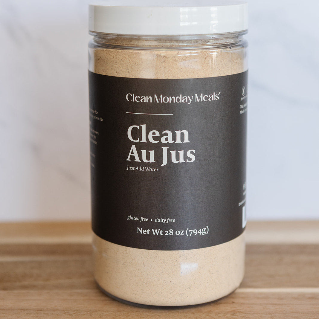 A clear plastic jar of Clean Monday Meals Clean Au Jus with a dark brown label and white lid, sitting on a wooden surface against a light marble background.
