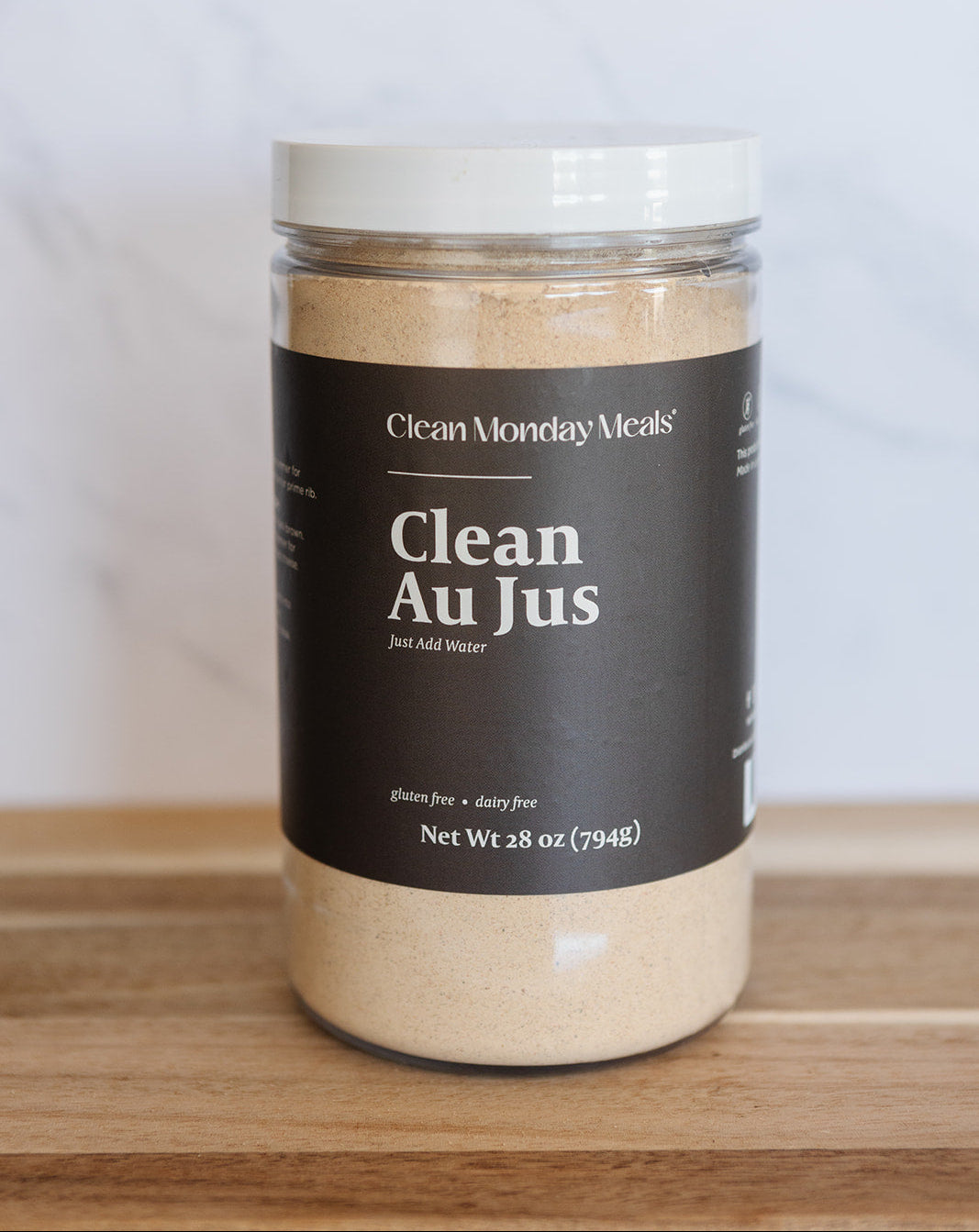 A clear plastic jar of Clean Monday Meals Clean Au Jus with a dark brown label and white lid, sitting on a wooden surface against a light marble background.