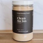 A clear plastic jar of Clean Monday Meals Clean Au Jus with a dark brown label and white lid, sitting on a wooden surface against a light marble background.