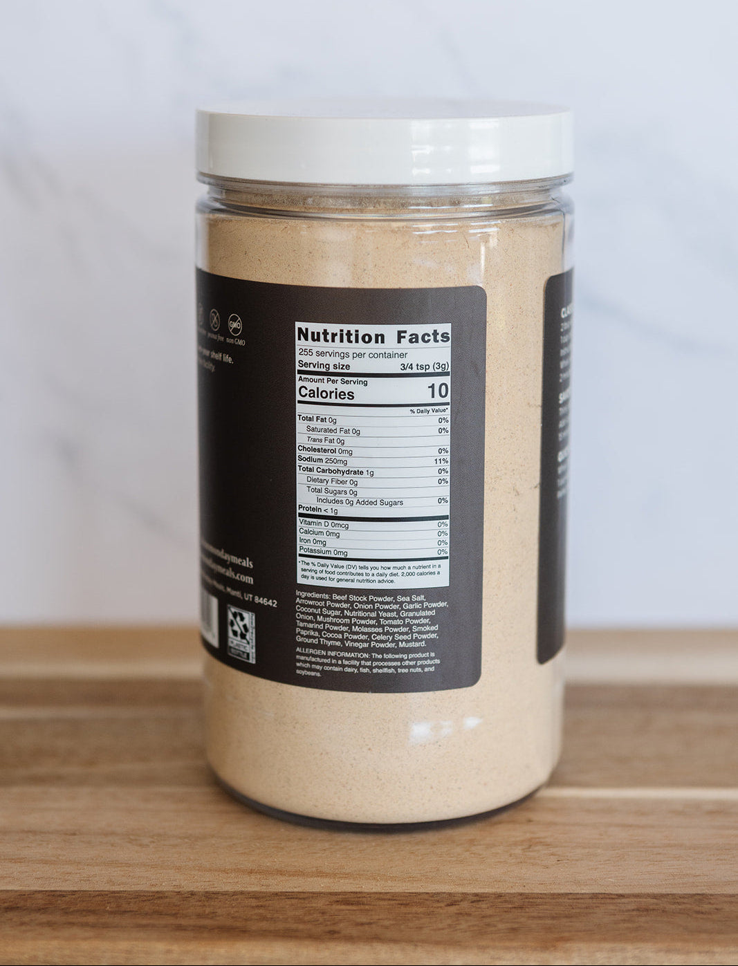 A clear plastic jar filled with a light beige powder, featuring a white lid and a black label showing the nutrition facts, set on a wooden surface against a light background.