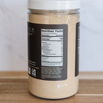 A clear plastic jar filled with a light beige powder, featuring a white lid and a black label showing the nutrition facts, set on a wooden surface against a light background.