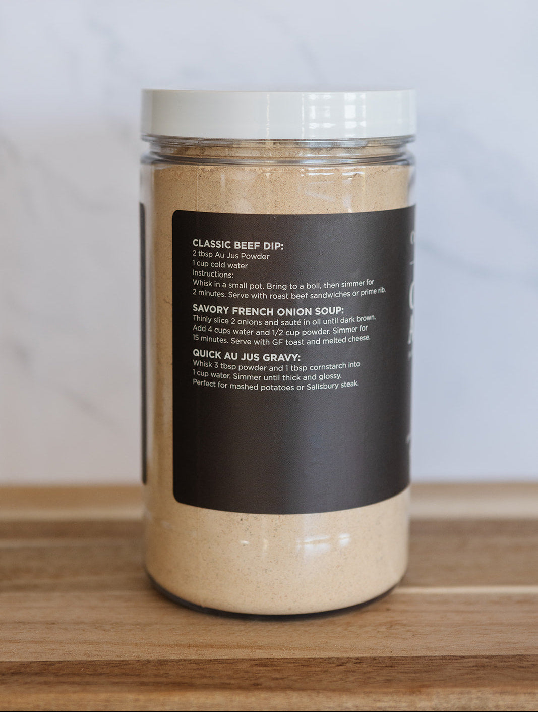 A clear plastic jar filled with a light tan beef dip powder, featuring a white lid and a black label with white text, set on a wooden surface against a light gray marble background.