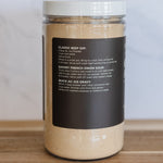 A clear plastic jar filled with a light tan beef dip powder, featuring a white lid and a black label with white text, set on a wooden surface against a light gray marble background.