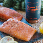 A bottle of Bayou Bold Seasoning by Clean Money Mealz stands on a rustic surface beside two salmon fillets, lemon slices, and green vegetables in a styled food scene.