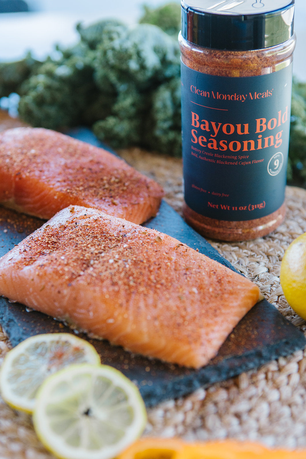 A bottle of Bayou Bold Seasoning by Clean Money Mealz stands on a rustic surface beside two salmon fillets, lemon slices, and green vegetables in a styled food scene.