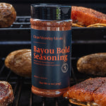 A bottle of Clean Monday's Bayou Bold Seasoning stands on a grill rack, surrounded by cooked meat and potatoes in a real outdoor cooking environment.