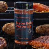 A bottle of Clean Monday's Bayou Bold Seasoning stands on a grill rack, surrounded by cooked meat and potatoes in a real outdoor cooking environment.