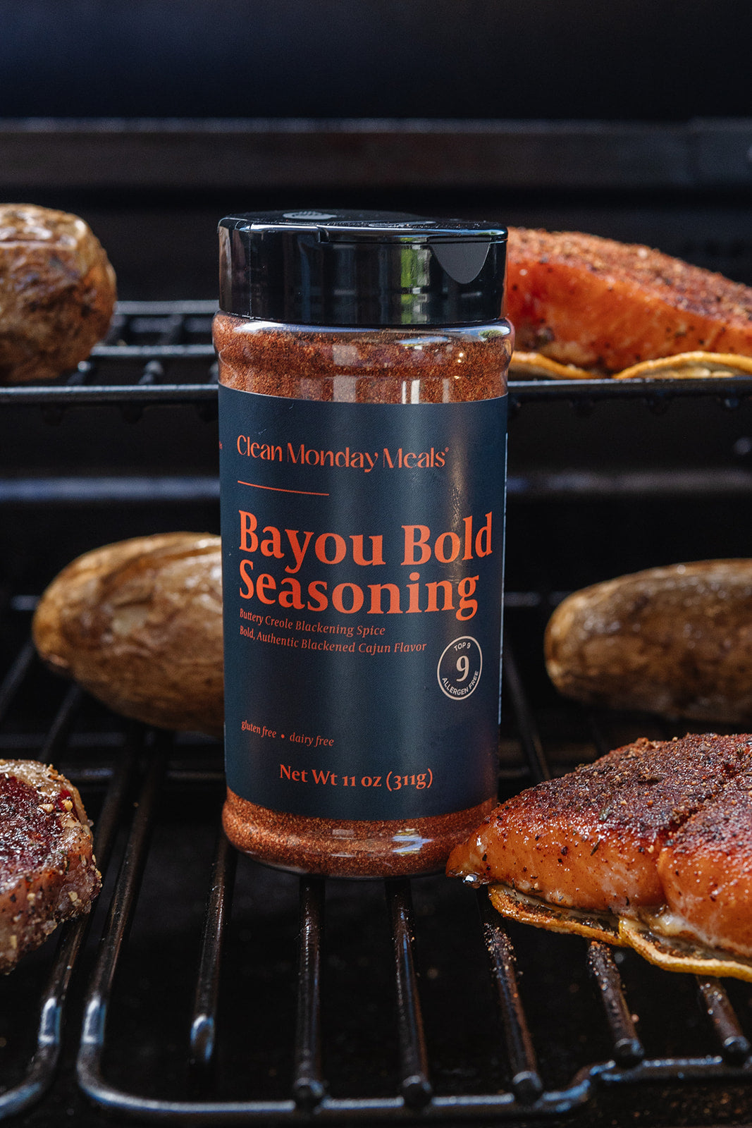 A bottle of Clean Monday's Bayou Bold Seasoning stands on a grill rack, surrounded by cooked meat and potatoes in a real outdoor cooking environment.