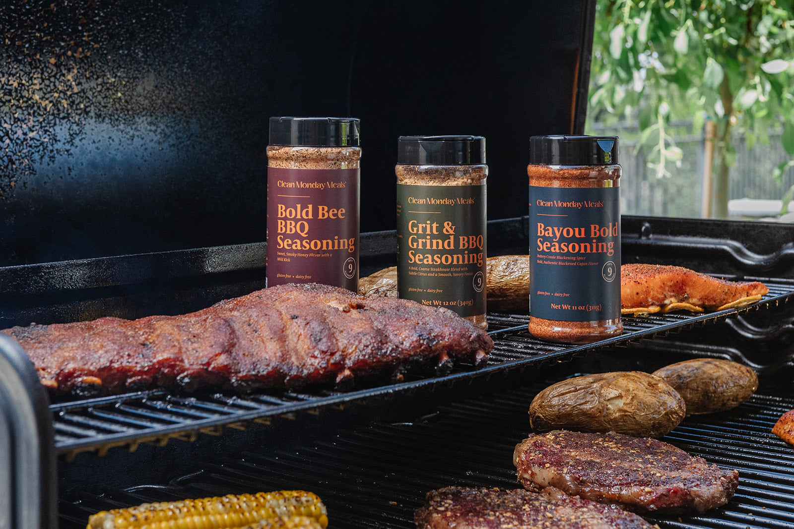 Three bottles of Charbrokers Wok seasonings standing on a black grill next to cooked ribs, potatoes, steak, and corn on a barbecue.