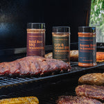 Three bottles of Charbrokers Wok seasonings standing on a black grill next to cooked ribs, potatoes, steak, and corn on a barbecue.