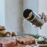 Person seasoning raw meat with a pepper grinder on a wooden cutting board.