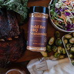 Bottle of Bold Bee BBQ seasoning on a wooden board with vegetables and salad.