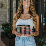 A smiling Janae Cox with long wavy blonde hair stands outdoors holding three jars of seasoning. She is wearing a white tank top and light blue denim shorts.