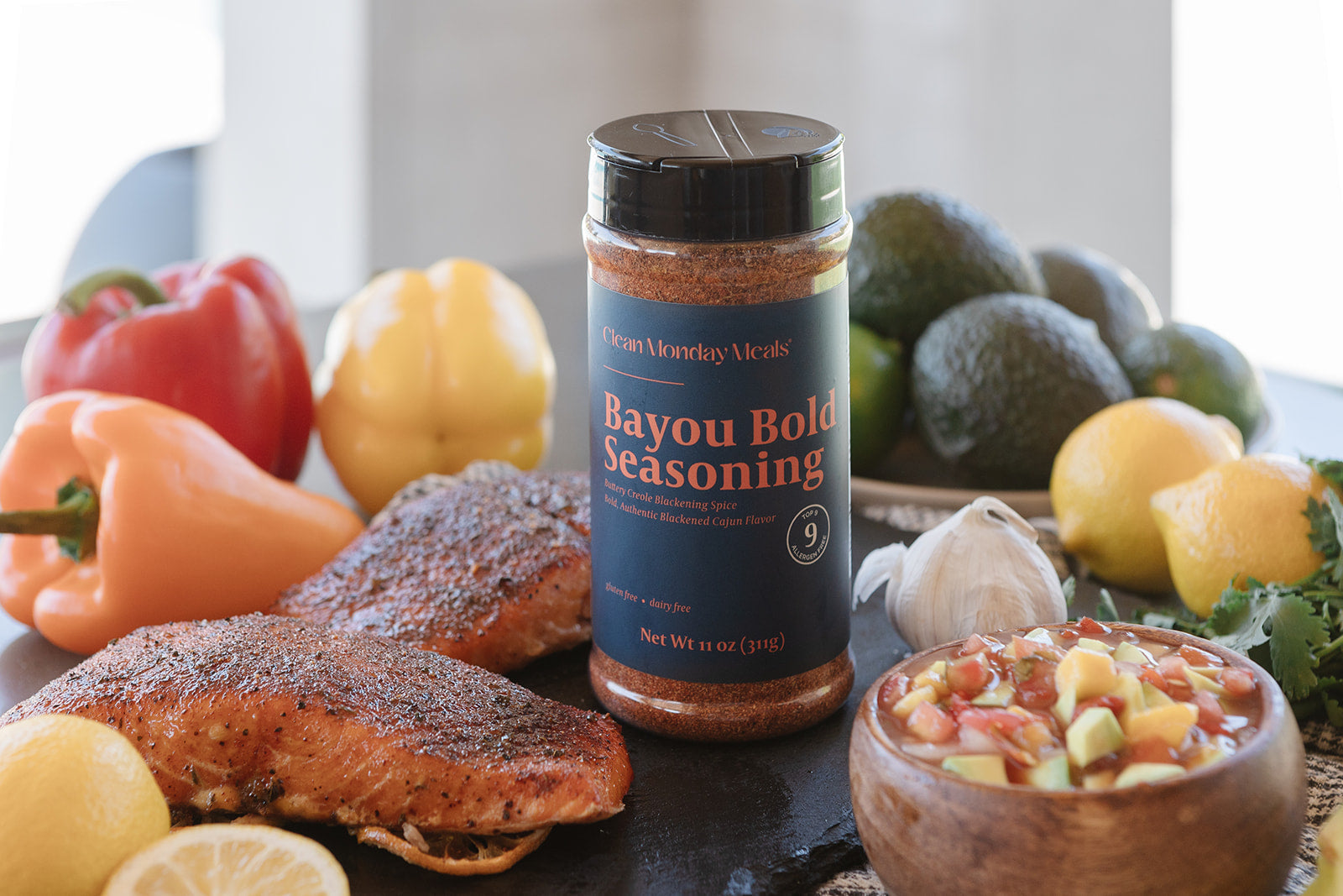 A bottle of Bayou Bold Seasoning stands on a dark slate board surrounded by cooked salmon fillets, lemons, bell peppers, avocados, garlic, and a bowl of salsa. The label reads 'Petite Kitchen Meals Bayou Bold Seasoning'.