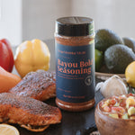A bottle of Bayou Bold Seasoning stands on a dark slate board surrounded by cooked salmon fillets, lemons, bell peppers, avocados, garlic, and a bowl of salsa. The label reads 'Petite Kitchen Meals Bayou Bold Seasoning'.