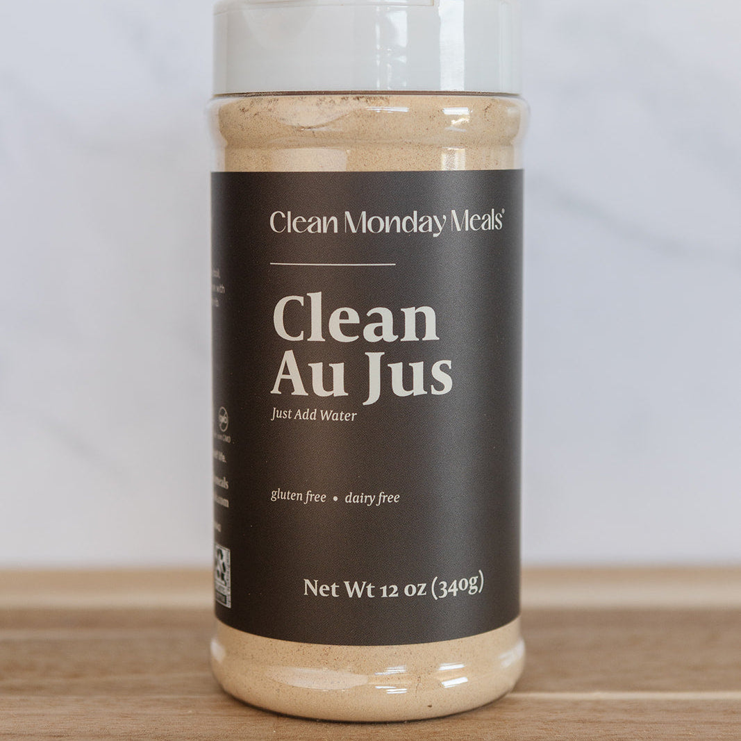 A 12 oz plastic jar of Clean Monday Meals Clean Au Jus on a wooden surface against a light gray marble background.