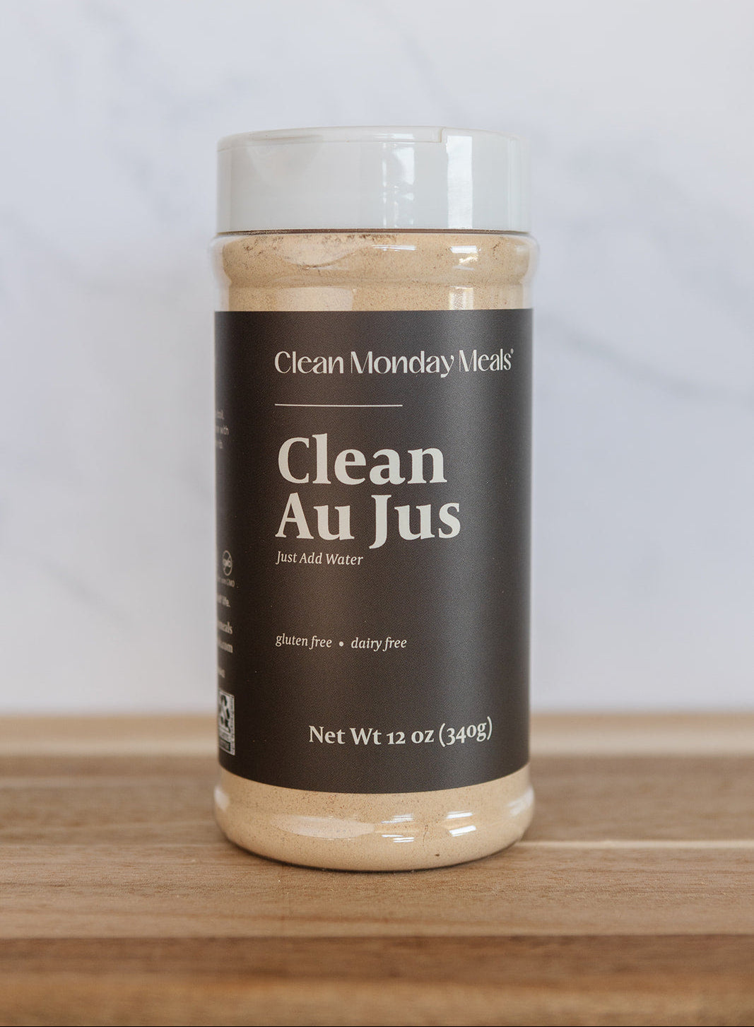 A 12 oz plastic jar of Clean Monday Meals Clean Au Jus on a wooden surface against a light gray marble background.