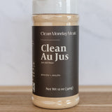 A 12 oz plastic jar of Clean Monday Meals Clean Au Jus on a wooden surface against a light gray marble background.