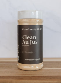 A 12 oz plastic jar of Clean Monday Meals Clean Au Jus on a wooden surface against a light gray marble background.