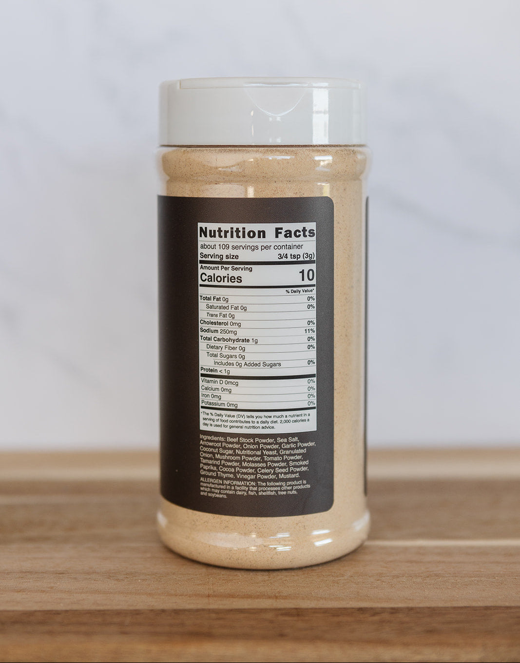 A clear plastic spice jar with a white lid, showing a beige powder inside and a black nutrition facts label on the front, set on a wooden surface against a light marble background.