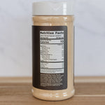 A clear plastic spice jar with a white lid, showing a beige powder inside and a black nutrition facts label on the front, set on a wooden surface against a light marble background.