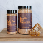 Two jars of Bold Bee BBQ Seasoning on a wooden surface with honeycomb and honey.
