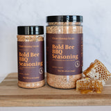 Two jars of Bold Bee BBQ Seasoning on a wooden surface with honeycomb and honey.