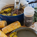 A close-up lifestyle shot of a cooking scene featuring a blue enameled cast iron pot with a hand using metal tongs to serve food. A jar of 'Clean Au Jus' beef seasoning is prominently displayed in the foreground, alongside corn on the cob, green peppers, and a dark plate.
