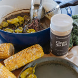 A close-up lifestyle shot of a cooking scene featuring a blue enameled cast iron pot with a hand using metal tongs to serve food. A jar of 'Clean Au Jus' beef seasoning is prominently displayed in the foreground, alongside corn on the cob, green peppers, and a dark plate.