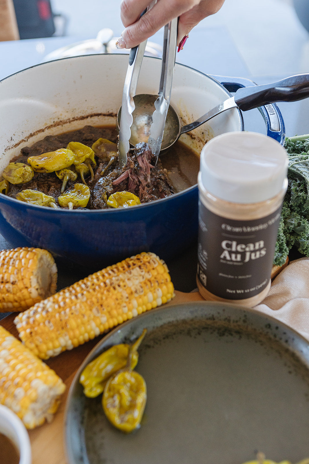 A close-up lifestyle shot of a cooking scene featuring a blue enameled cast iron pot with a hand using metal tongs to serve food. A jar of 'Clean Au Jus' beef seasoning is prominently displayed in the foreground, alongside corn on the cob, green peppers, and a dark plate.