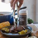 A close-up lifestyle shot of a hand using metal tongs to lift shredded pulled meat from a dark plate, with a jar labeled 'Clean Au Jus' in the background and cooked food items around the table.