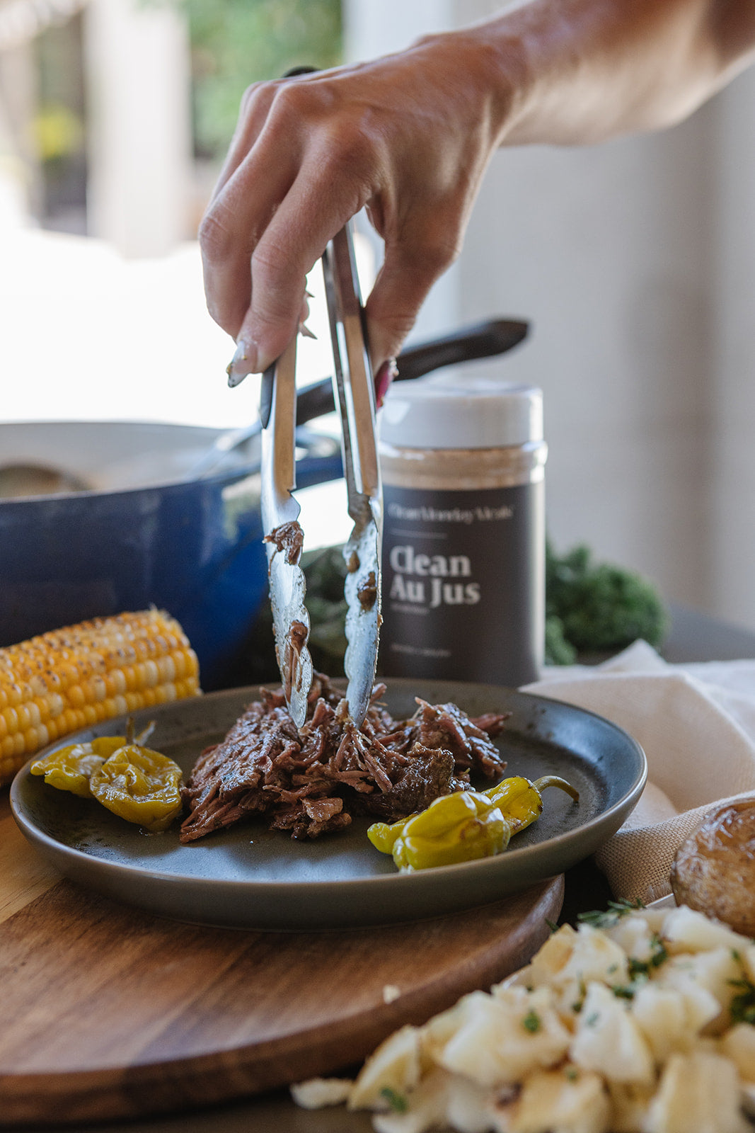 A close-up lifestyle shot of a hand using metal tongs to lift shredded pulled meat from a dark plate, with a jar labeled 'Clean Au Jus' in the background and cooked food items around the table.