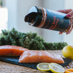 A hand sprinkles seasoning from a black shaker bottle onto raw salmon fillets on a dark slate board, with kale, lemons, and an orange in a kitchen setting.