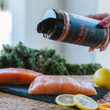 A hand sprinkles seasoning from a black shaker bottle onto raw salmon fillets on a dark slate board, with kale, lemons, and an orange in a kitchen setting.