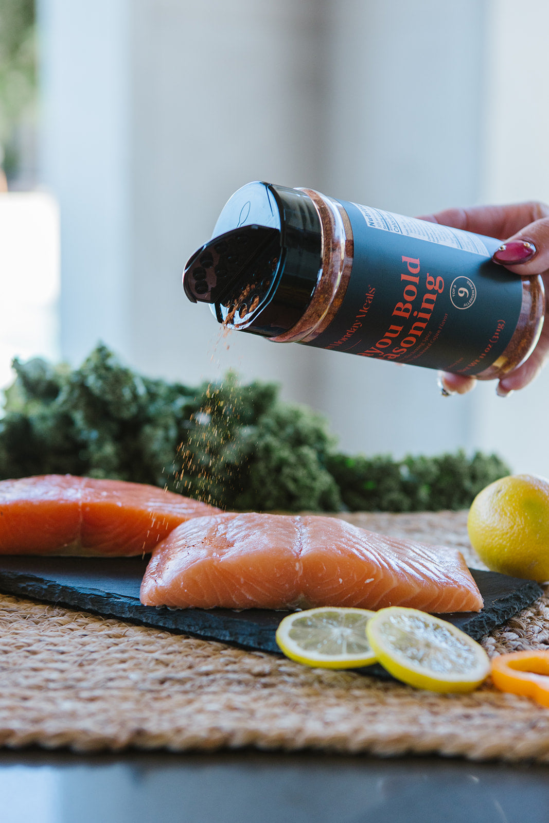 A hand sprinkles seasoning from a black shaker bottle onto raw salmon fillets on a dark slate board, with kale, lemons, and an orange in a kitchen setting.