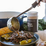 A close-up lifestyle shot of a hand using a ladle to pour sauce over shredded beef on a dark plate, with a jar of beef jus seasoning in the background and other cooked foods on a wooden board.
