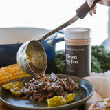 A close-up lifestyle shot of a hand using a ladle to pour sauce over shredded beef on a dark plate, with a jar of beef jus seasoning in the background and other cooked foods on a wooden board.
