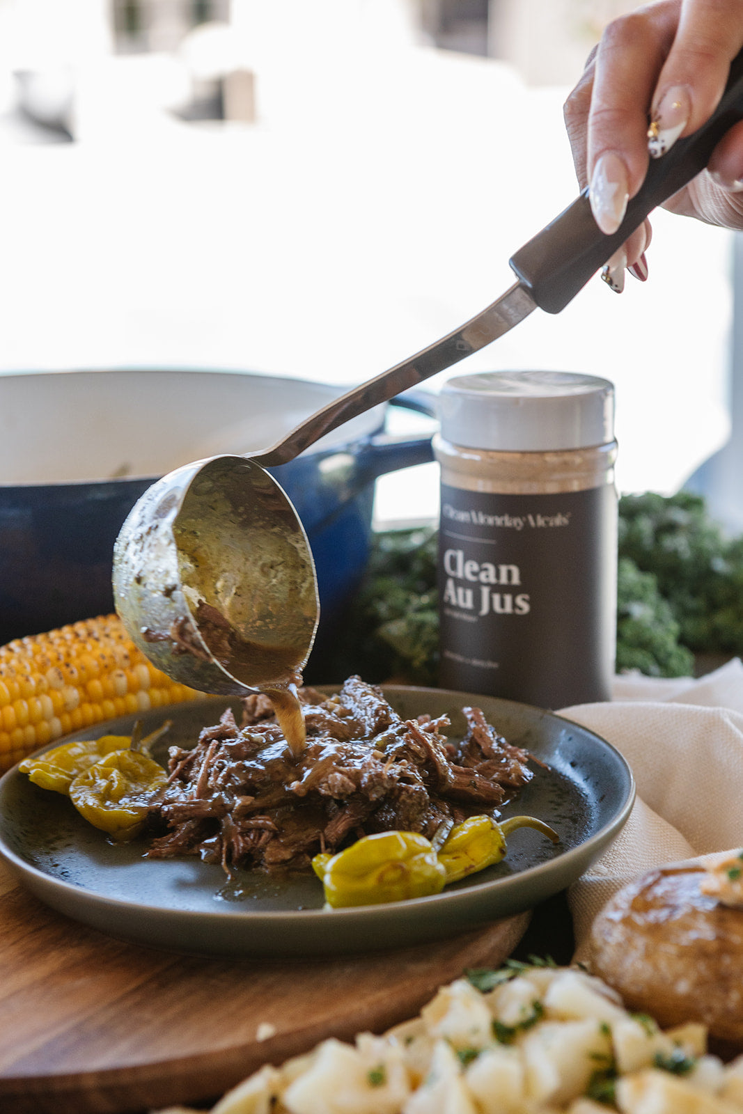 A close-up lifestyle shot of a hand using a ladle to pour sauce over shredded beef on a dark plate, with a jar of beef jus seasoning in the background and other cooked foods on a wooden board.