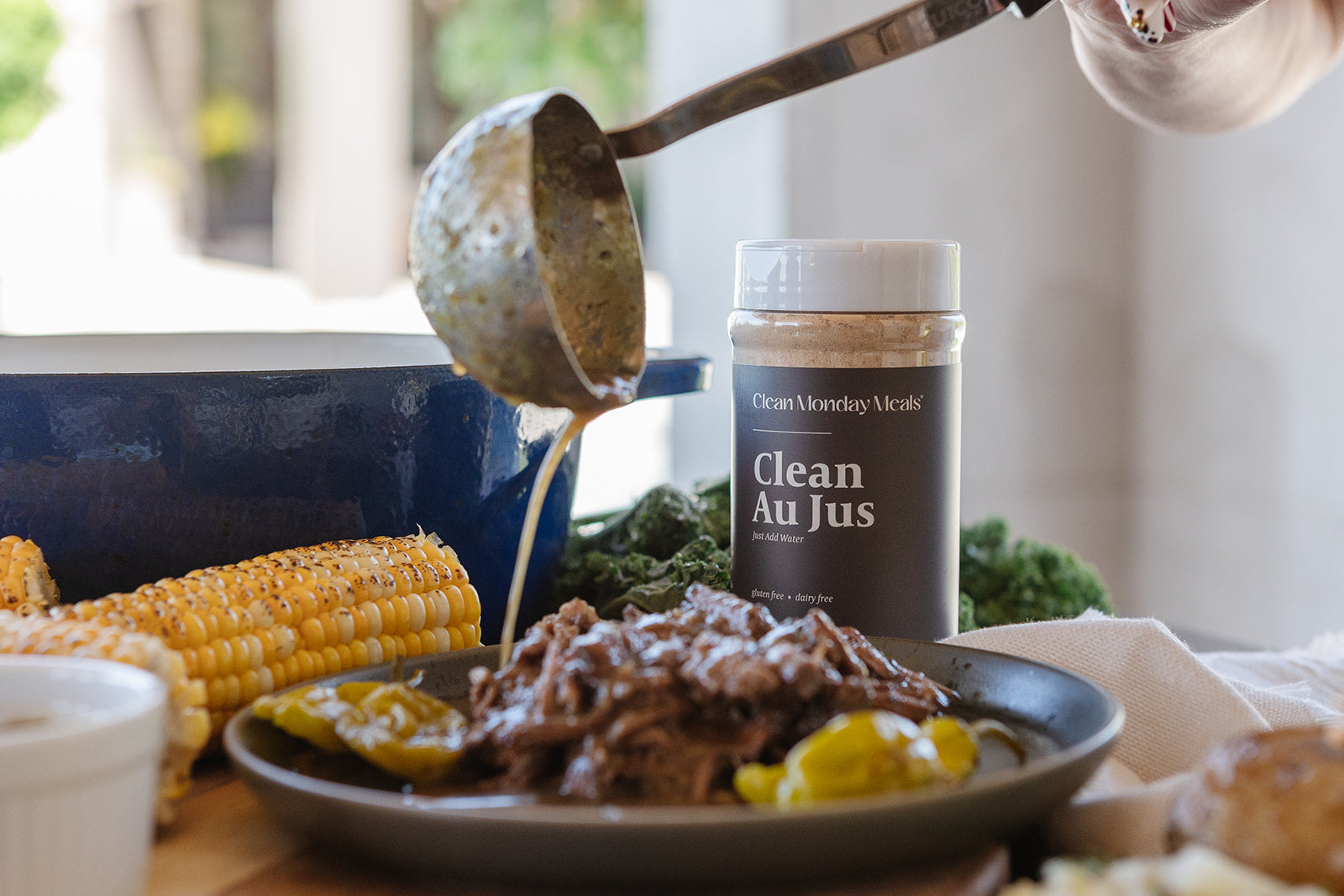 A jar of Clean Monday Meals Clean Au Jus seasoning stands on a table beside a plated cooked meat dish, corn on the cob, and a ladle pouring sauce into the plate in a real kitchen setting.
