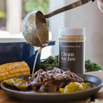 A jar of Clean Monday Meals Clean Au Jus seasoning stands on a table beside a plated cooked meat dish, corn on the cob, and a ladle pouring sauce into the plate in a real kitchen setting.